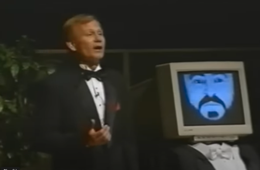Dr. Ingo titze stands in a tuxedo on a stage next to a computer screen that features an image resembling Luciano Pavarotti's face.